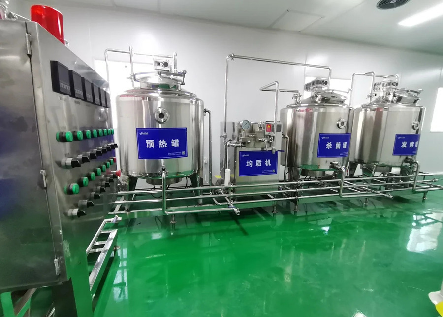 An industrial view of the complete, integrated dairy processing line, featuring a large control cabinet, followed by a preheating tank, a small homogenizer unit, a sterilization tank, and a fermentation tank, all connected by sanitary piping on a clean green floor. 
